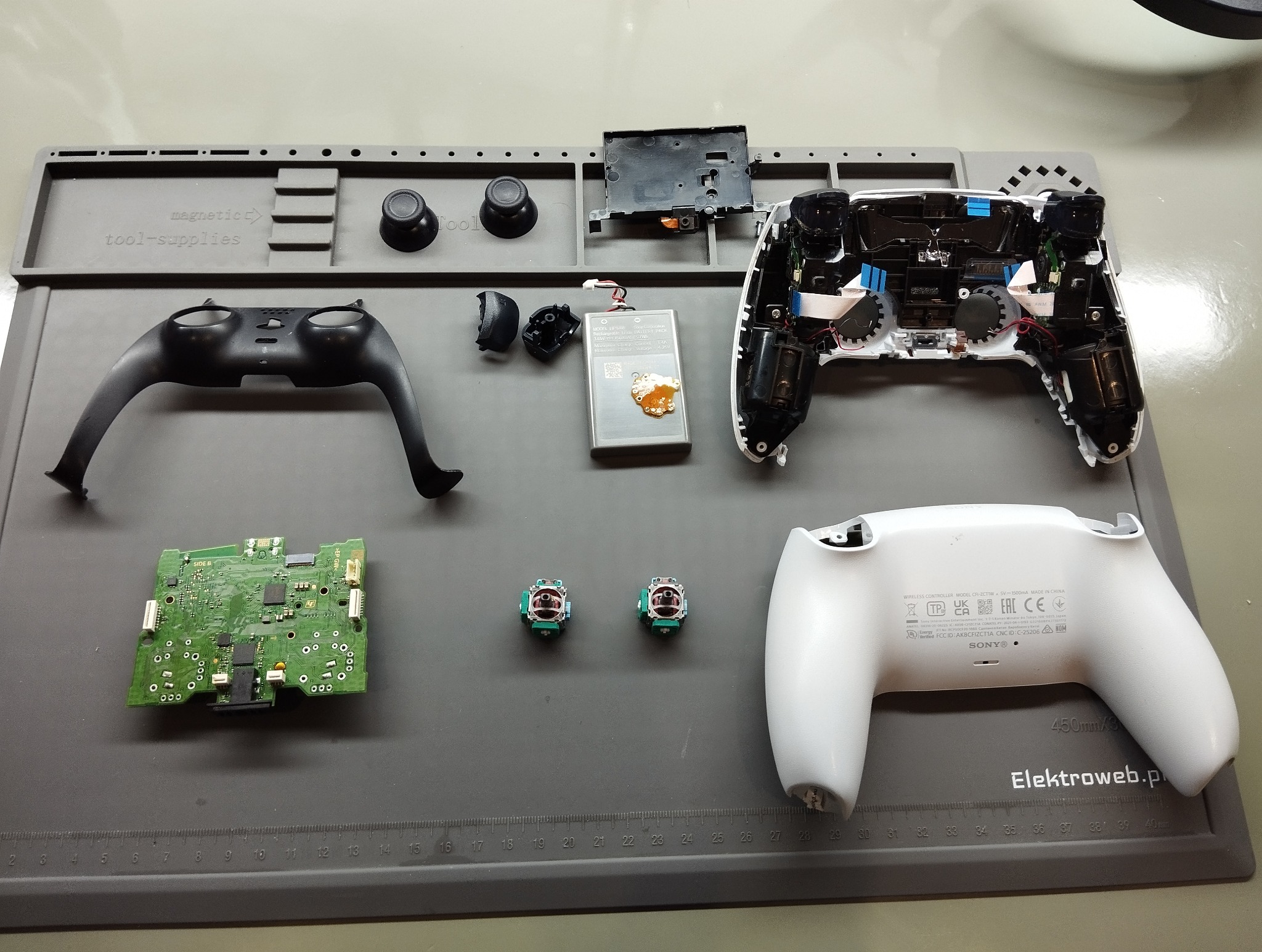 Rozłożony kontroler DualSense od PlayStation 5 na macie serwisowej. Widoczne elementy: obudowa, bateria, płyta główna, analogi. Precyzyjna naprawa sprzętu elektronicznego.