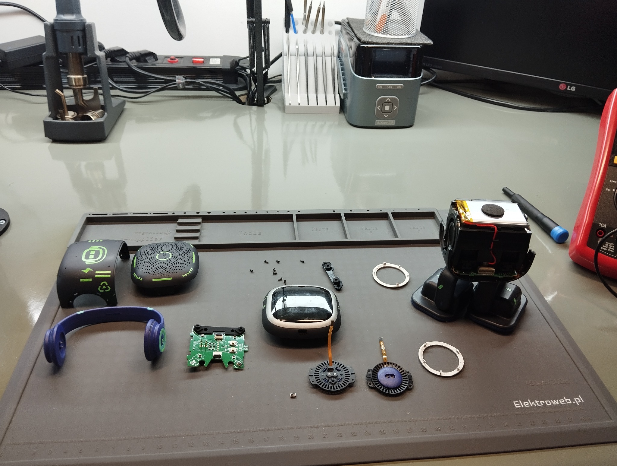Rozłożone na stole słuchawki, kamera sportowa i inne akcesoria elektroniczne w trakcie naprawy. Widoczne narzędzia i mata serwisowa.