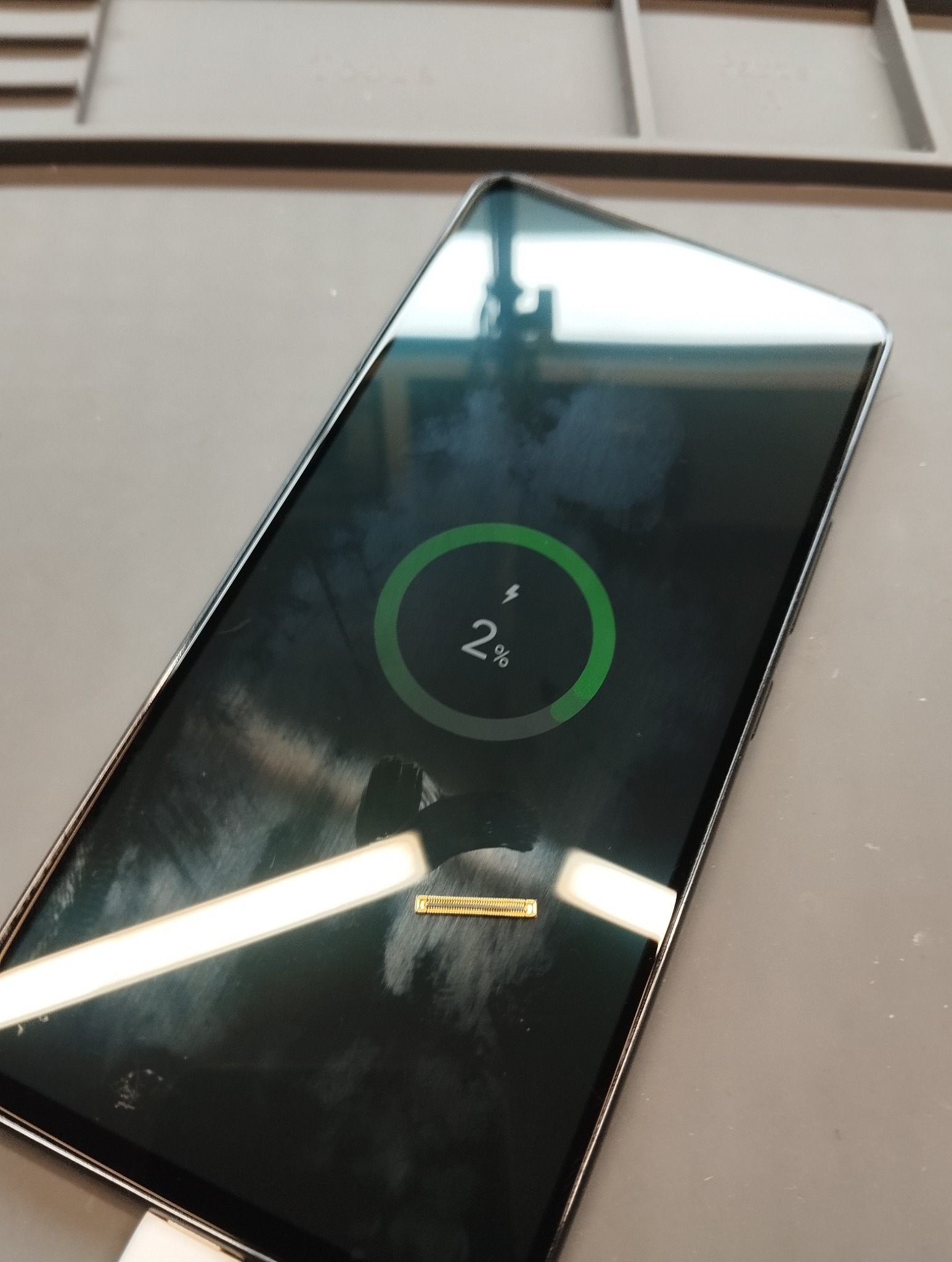 Smartfon z ekranem wskazującym 2% naładowania baterii, widoczny port ładowania. Sprzęt leży na szarej macie serwisowej.