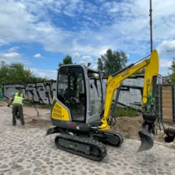 Żółto-szara mini koparka Wacker Neuson ET18 na brukowanej powierzchni, pracownik w kamizelce odblaskowej zamiata obok, w tle graffiti na metalowym ogrodzeniu i błękitne niebo z chmurami.