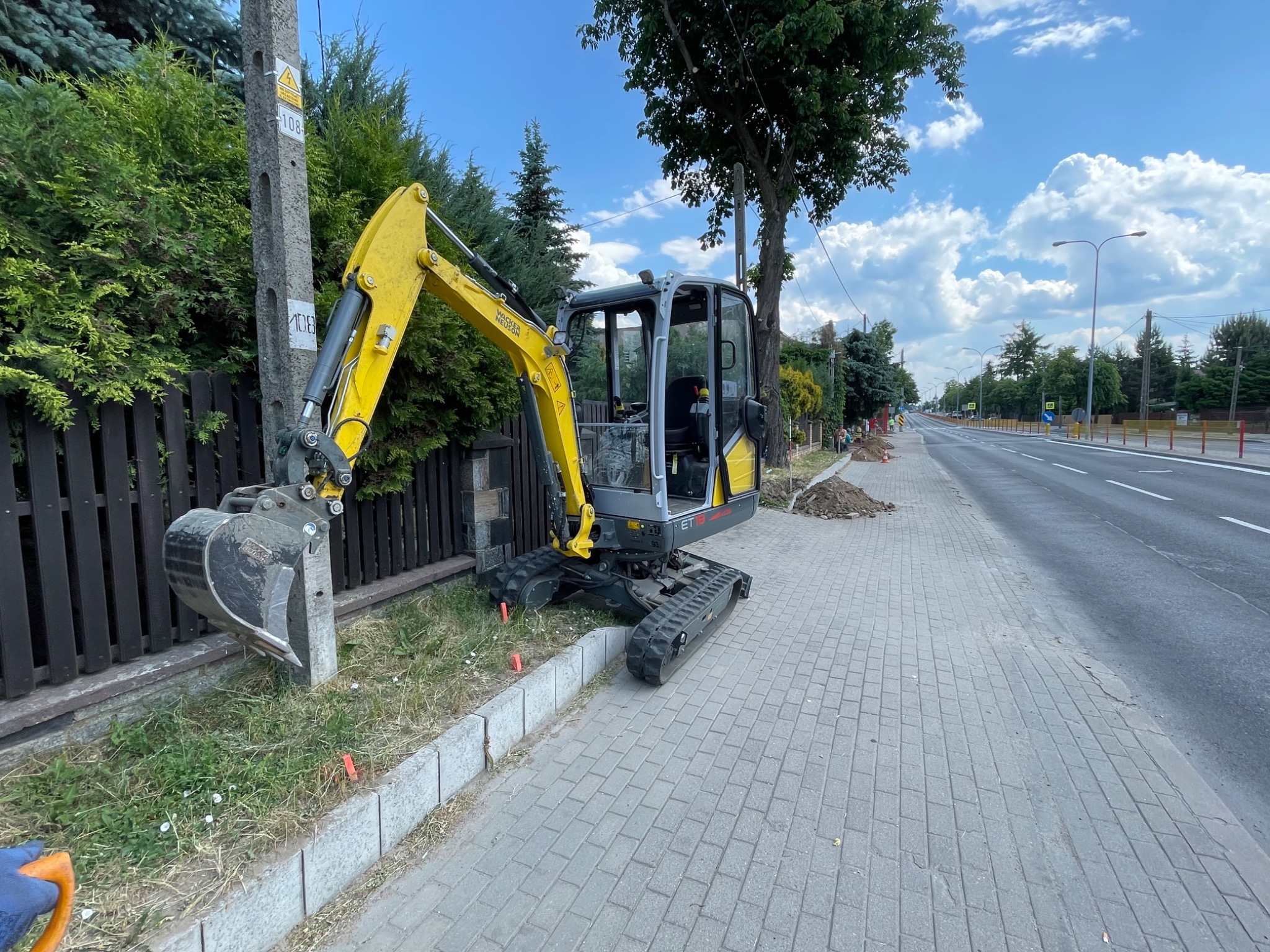 Żółto-szara mini koparka Wacker Neuson ETT18 na chodniku z kostki brukowej, obok ogrodzenia i trawnika, w tle ulica i drzewa.