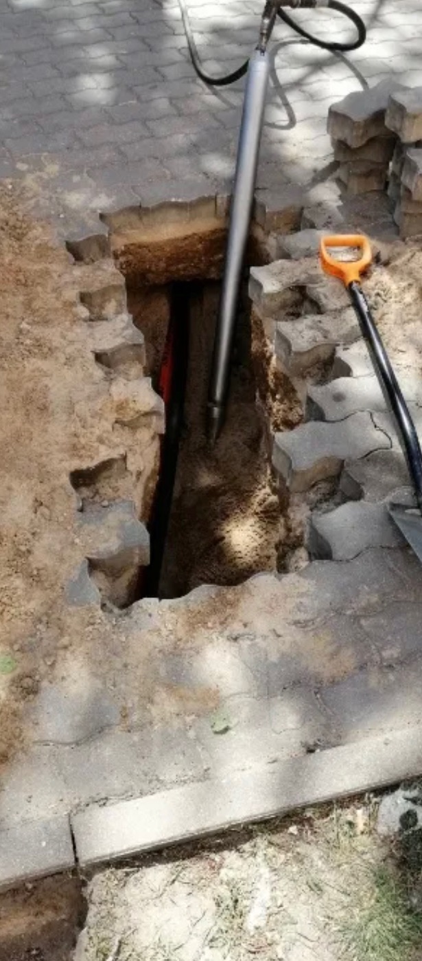 Wąski wykop w kostce brukowej, wewnątrz widoczne rury, obok leży łopata, a w ziemię wbita jest sonda geotechniczna.