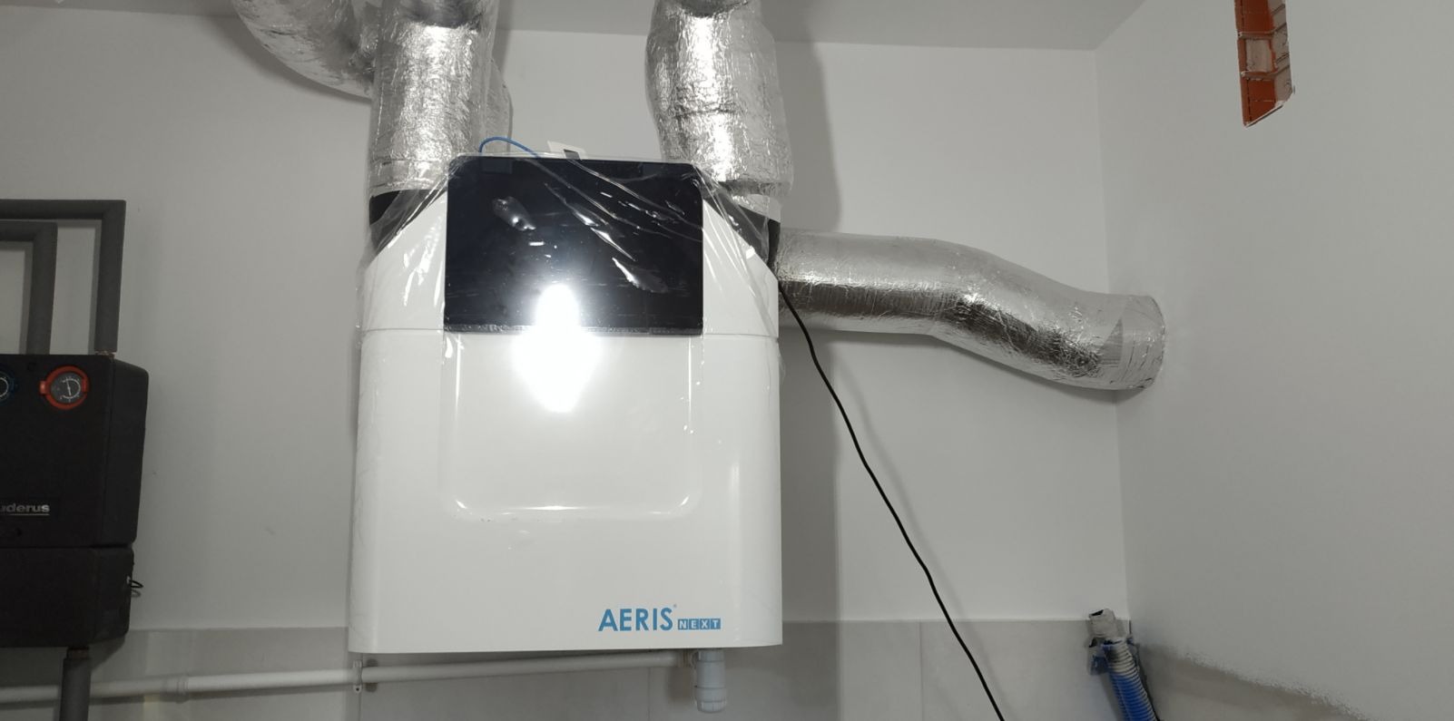 Biała jednostka centralna rekuperacji AERIS NEXT z podłączonymi, izolowanymi przewodami wentylacyjnymi, umieszczona w pomieszczeniu technicznym podczas montażu, wciąż zabezpieczona folią ochronną.
