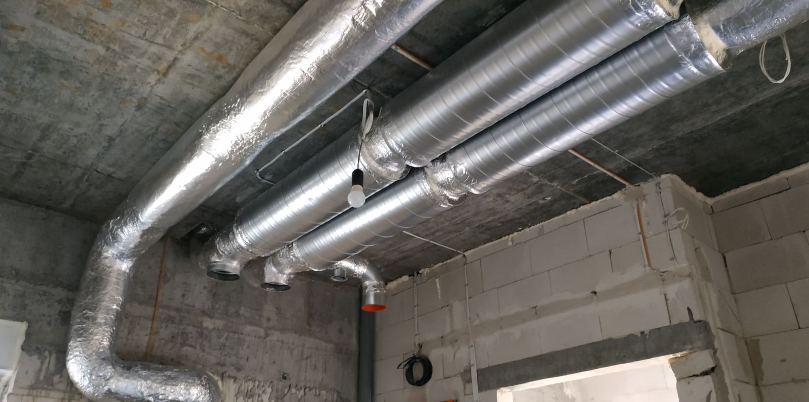 Montaż izolowanych i nieizolowanych przewodów wentylacyjnych pod betonowym stropem w surowym wnętrzu budynku.