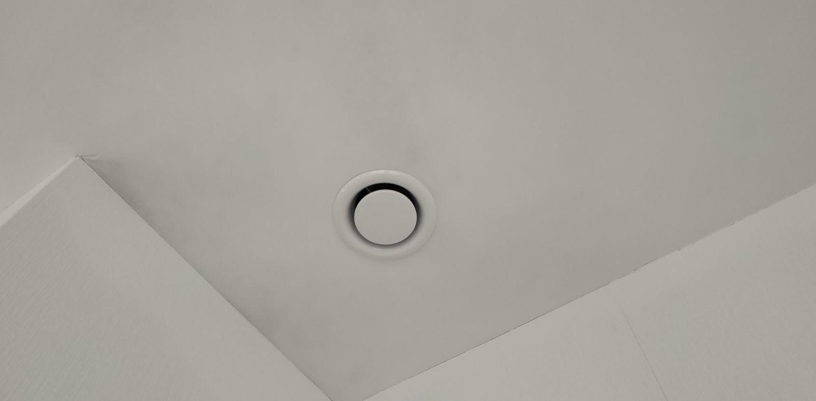 Okrągły, biały nawiewnik sufitowy zamontowany w narożniku pomieszczenia o jasnych ścianach.