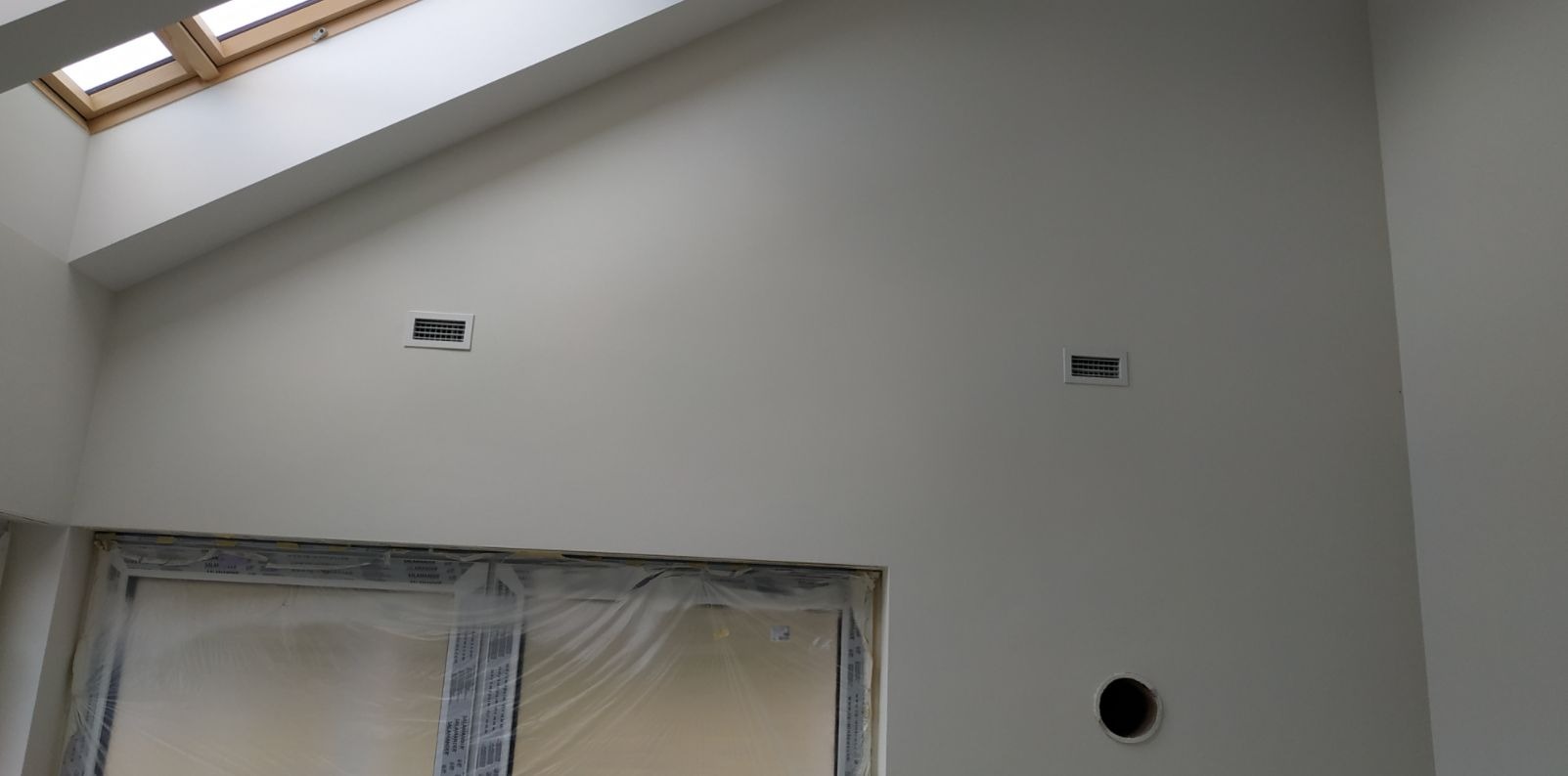 Dwa prostokątne nawiewniki wentylacyjne zamontowane na jasnej ścianie pod skośnym sufitem z oknem dachowym, widoczne również okno zabezpieczone folią i otwór w ścianie.