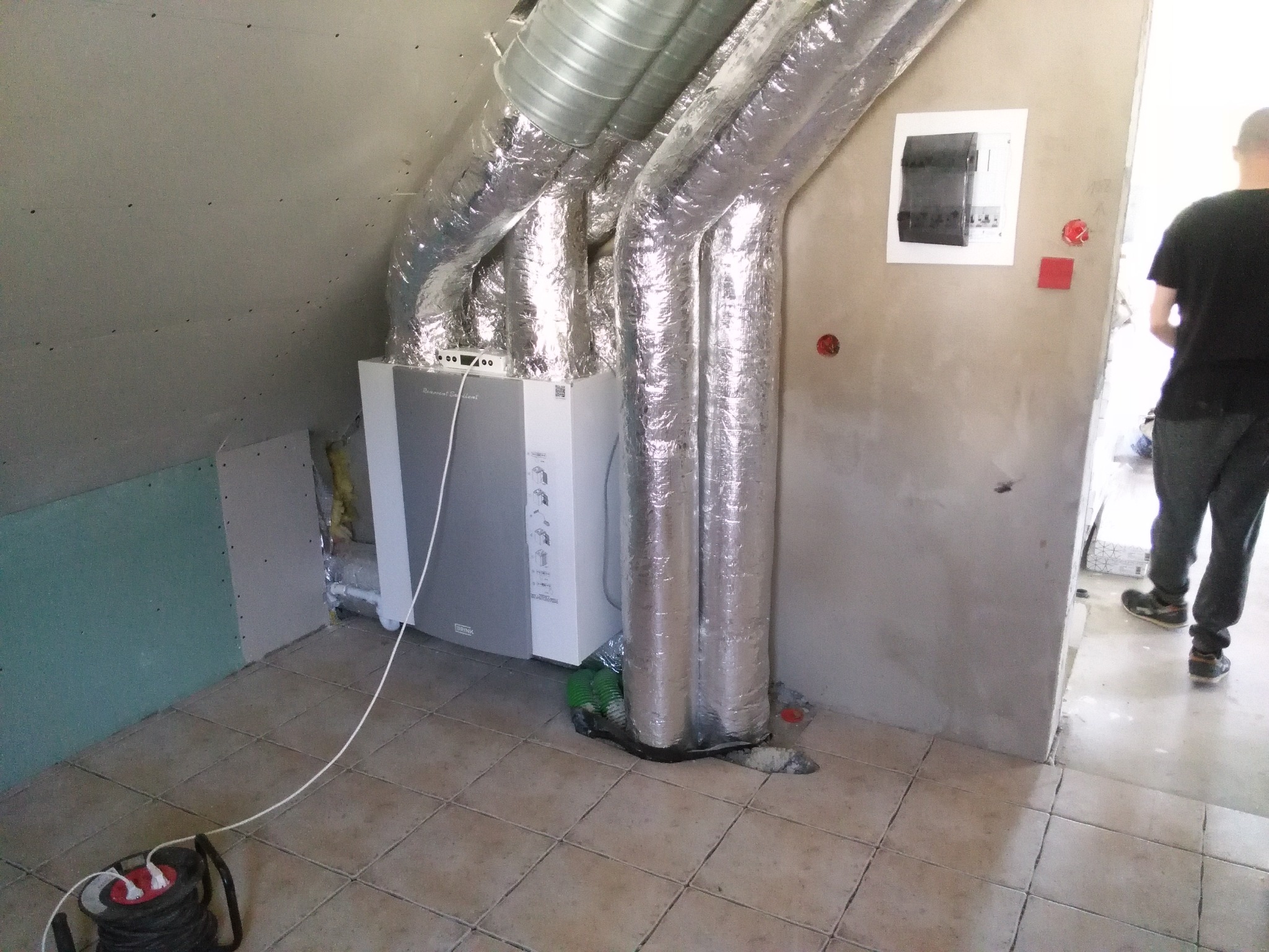 Szary rekuperator Brink zamontowany w pomieszczeniu z widocznymi przewodami wentylacyjnymi w srebrnej izolacji, podłączony do przedłużacza na podłodze z płytek.