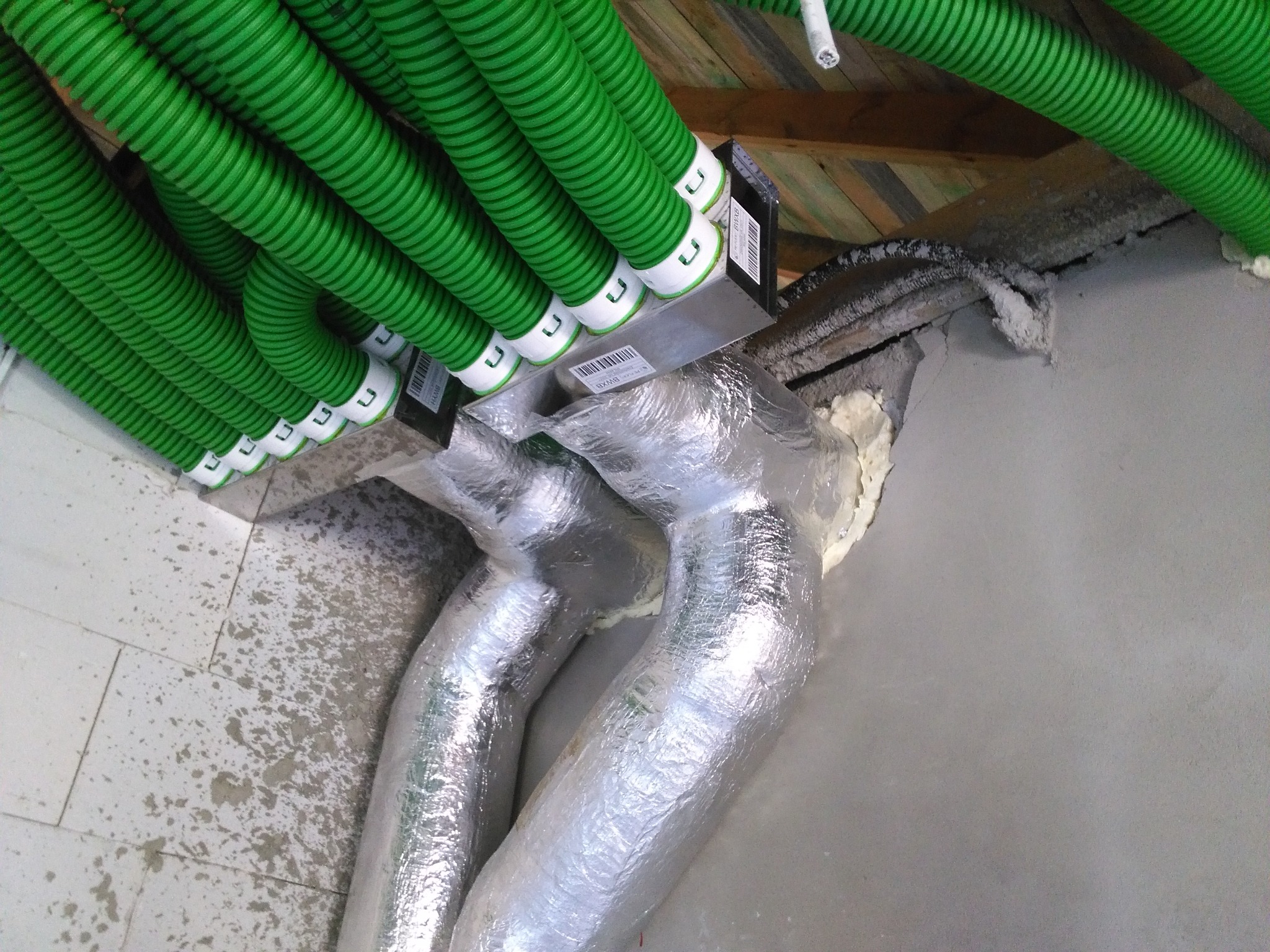 Instalacja systemu wentylacji: widok z dołu na zielone, karbowane rury wentylacyjne przymocowane do sufitu, przechodzące w srebrne, izolowane przewody. Widoczne elementy montażowe i pianka...