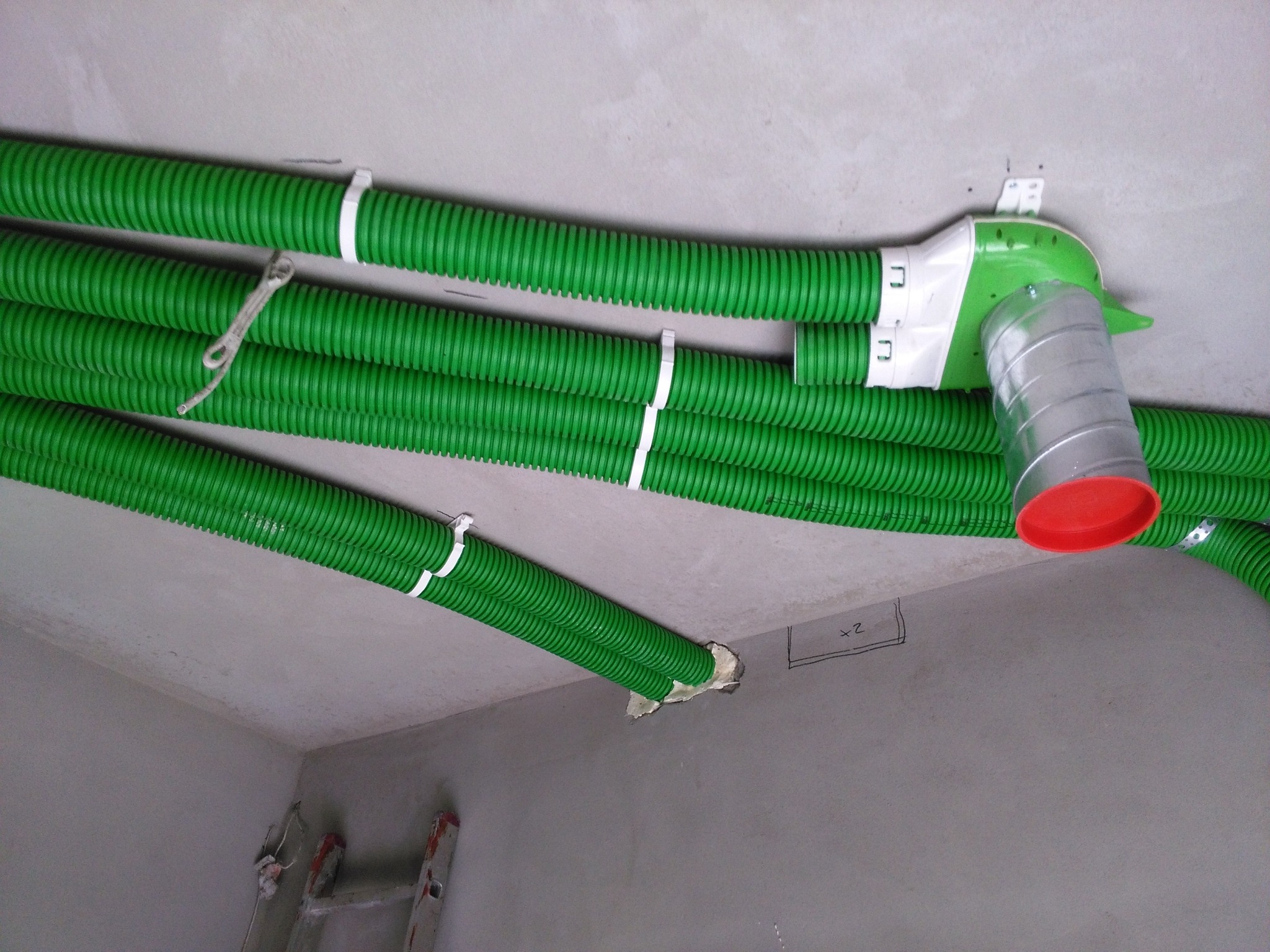 Montaż systemu wentylacji: zielone, karbowane rury wentylacyjne przymocowane do sufitu za pomocą białych opasek, widoczny element łączący z czerwonym zakończeniem, w tle drabina.