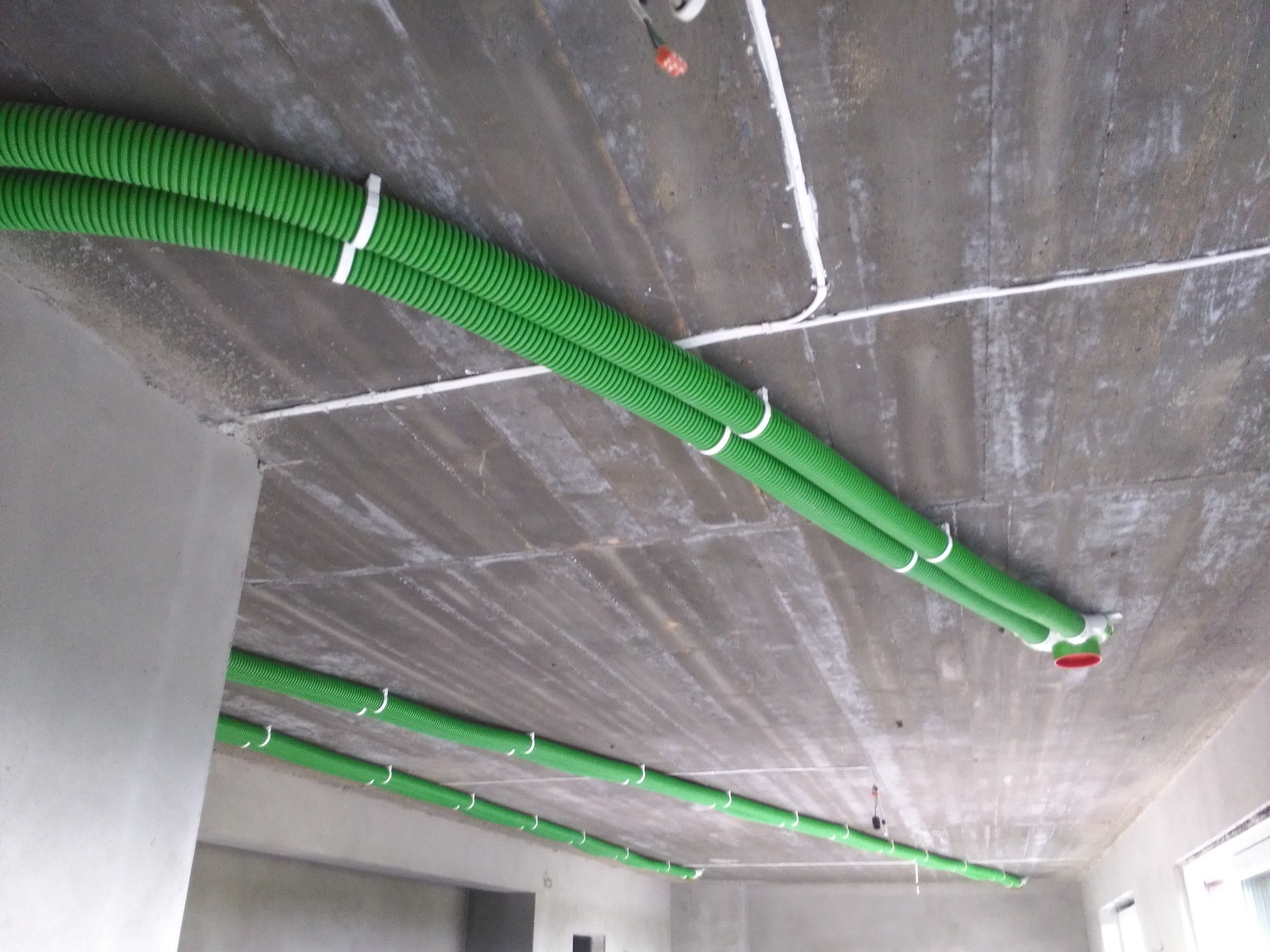 Zielone, karbowane rury wentylacyjne przymocowane do sufitu za pomocą białych opasek zaciskowych w surowym stanie deweloperskim.