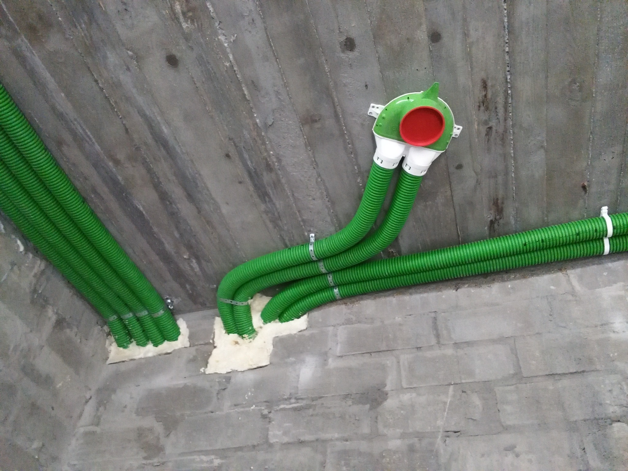 Montaż instalacji rekuperacji: zielone, karbowane rury wentylacyjne przymocowane do surowego betonowego sufitu i ściany z cegły, uszczelnione pianką montażową, z widocznym anemostatem w kolorze...