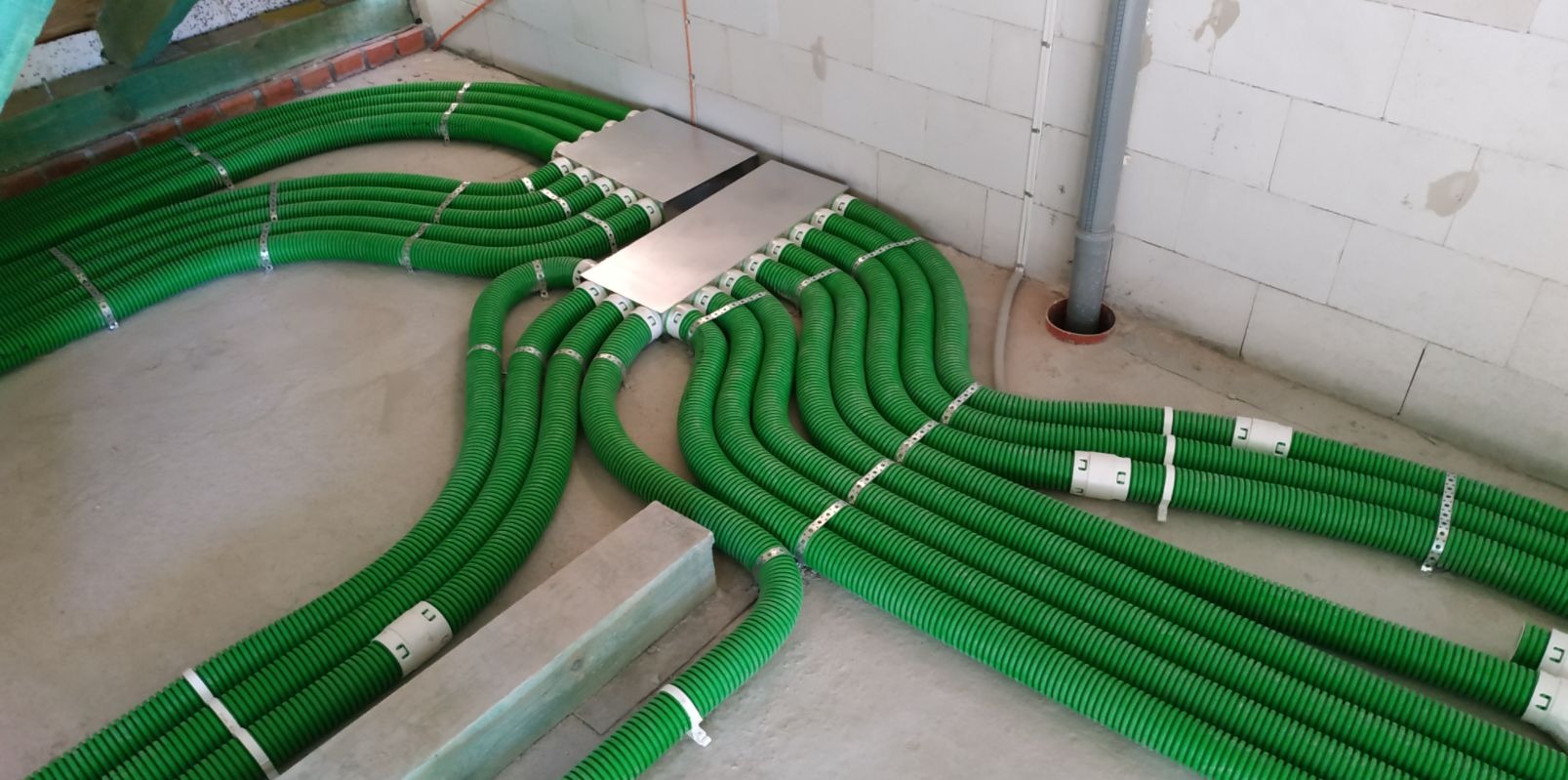 Rozprowadzenie zielonych rur wentylacyjnych z metalowymi obejmami w surowym wnętrzu, widoczne dwie metalowe skrzynki rozdzielcze.