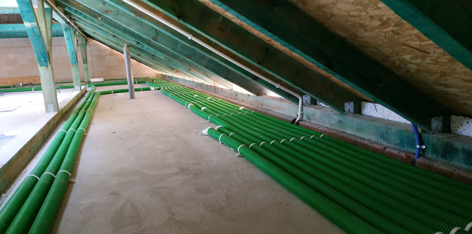 Zielone, karbowane rury wentylacyjne ułożone równolegle w surowym stanie poddasza z drewnianą konstrukcją dachu i betonową podłogą.