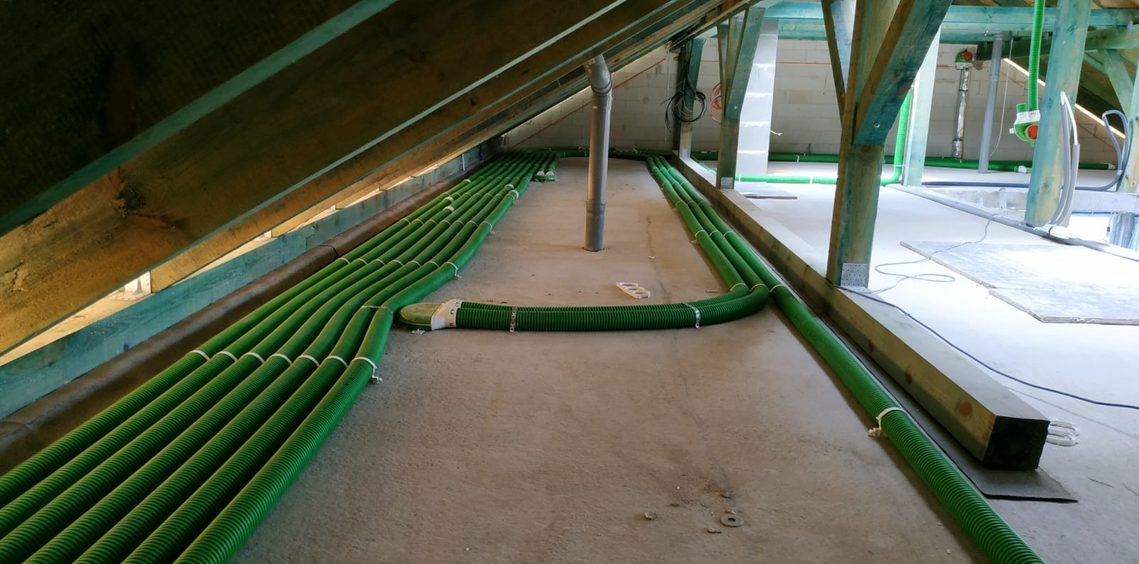 Zielone, karbowane rury wentylacyjne ułożone równolegle na poddaszu, przymocowane białymi opaskami, widoczne drewniane elementy konstrukcji dachu i pionowy przewód wentylacyjny.