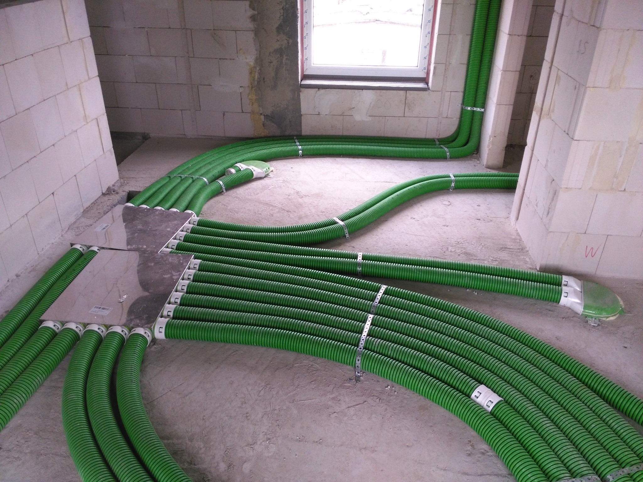 Zielone, karbowane rury wentylacyjne rozprowadzone po podłodze w surowym stanie, przymocowane metalowymi obejmami do podłoża, prowadzące do otworów w ścianie z pustaków.