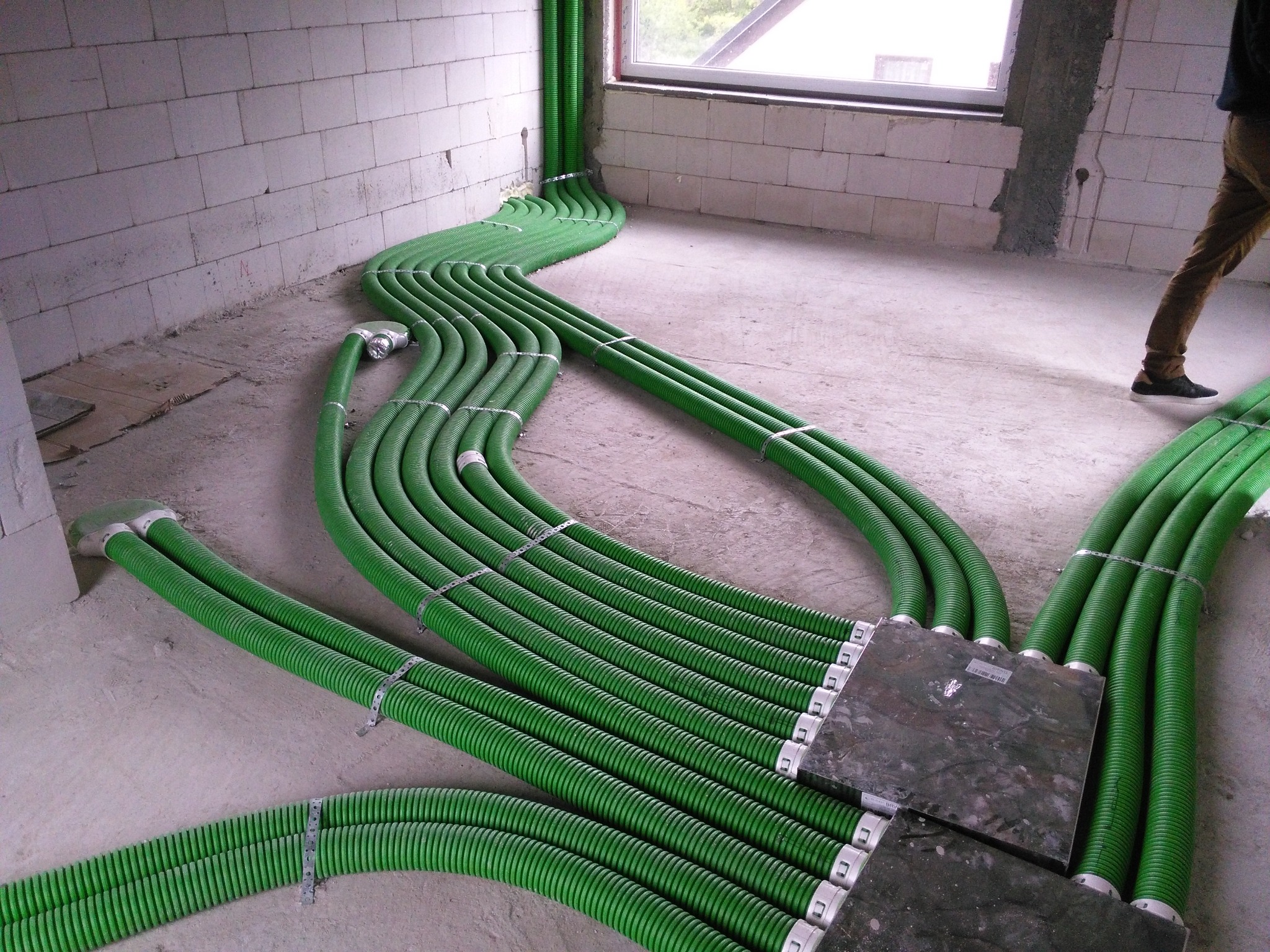 Wnętrze w trakcie instalacji systemu wentylacyjnego z widocznymi zielonymi, karbowanymi rurami rozprowadzonymi po podłodze w surowym stanie, mocowanymi metalowymi obejmami.