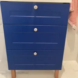 Niebieska szafka łazienkowa z trzema szufladami i ceramicznymi uchwytami na tle marmurowej ściany i podłogi.