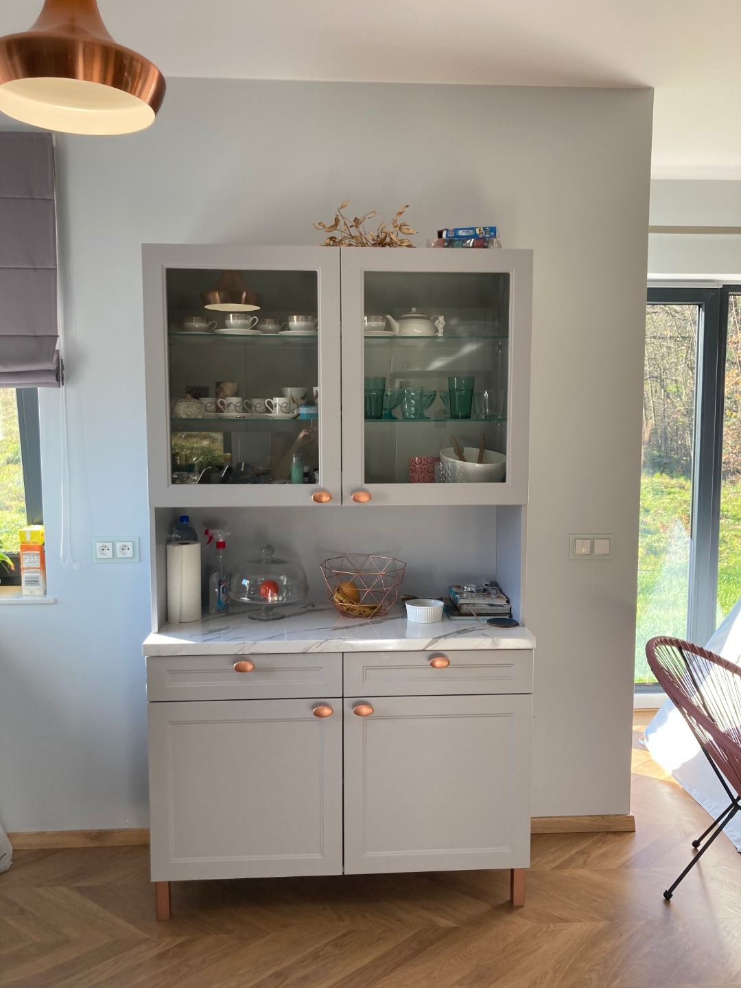 Szara witryna kuchenna z przeszklonymi drzwiczkami, marmurowym blatem i miedzianymi uchwytami, na blacie dekoracje i owoce pod kloszem, podłoga w jodełkę.