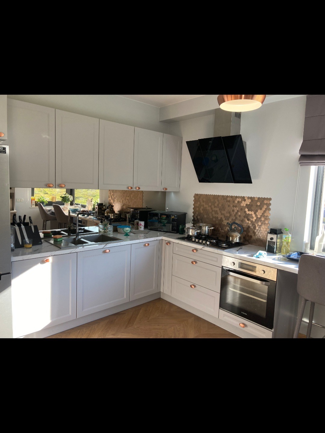 Jasna kuchnia z białymi frontami szafek i miedzianymi uchwytami, marmurowy blat, heksagonalna mozaika na ścianie, widok na ogród w odbiciu lustra.