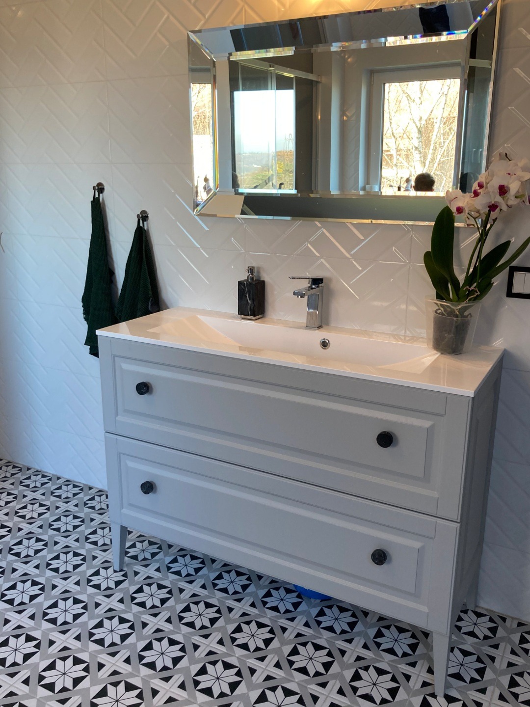 Szara szafka łazienkowa z dwiema szufladami pod umywalką, czarne gałki, białe płytki ułożone w jodełkę na ścianie, lustro z fazowanymi krawędziami, wzorzysta podłoga w czarno-białe gwiazdy.