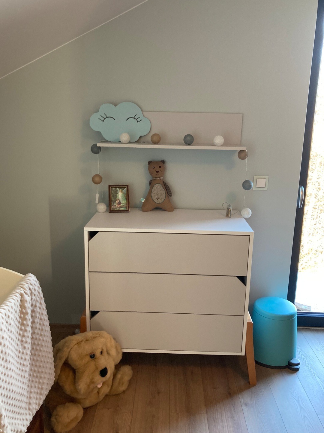 Jasnoszara komoda z trzema szufladami w pokoju dziecięcym, na blacie lampka w kształcie chmurki, pluszowy miś i girlanda świetlna, obok stoi błękitny kosz na śmieci.