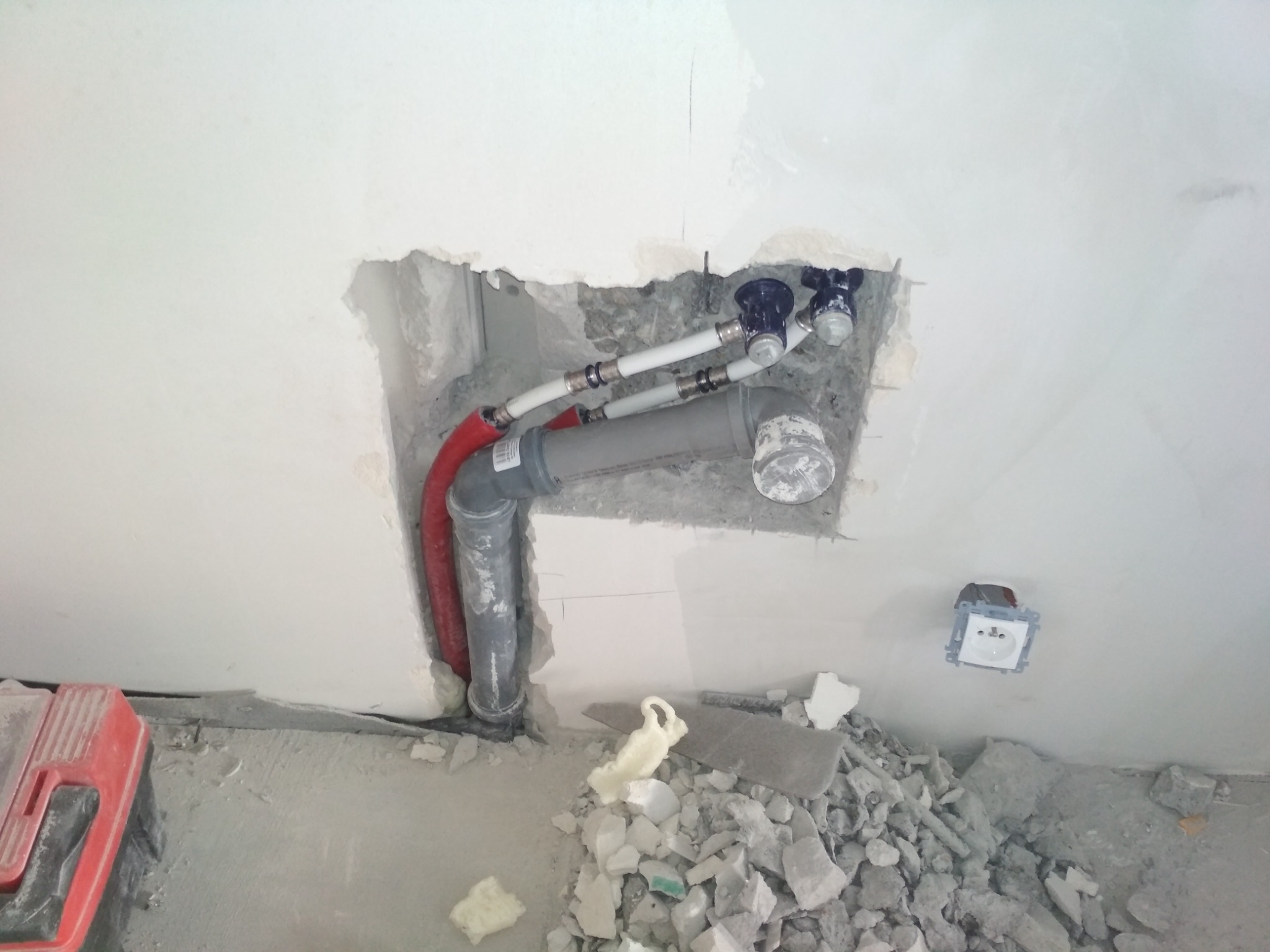 Fragment ściany z odsłoniętymi rurami hydraulicznymi i zaworami, widoczne gruz i resztki materiałów budowlanych na podłodze, gniazdko elektryczne w tle.