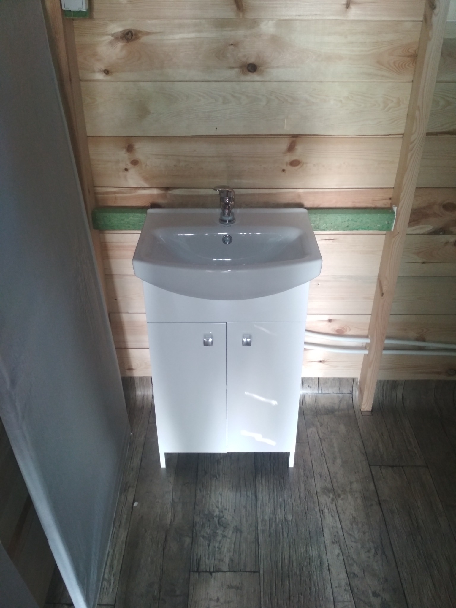 Nowa, biała umywalka z szafką zamontowana w łazience wykończonej drewnem w Gorzowie Wielkopolskim. Widoczne rury doprowadzające wodę.