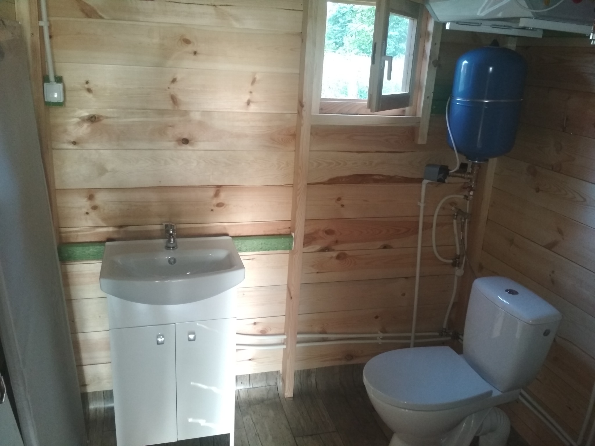 Wnętrze łazienki wykończonej drewnem z białą umywalką z szafką, toaletą i niebieskim zbiornikiem ciśnieniowym przy oknie.