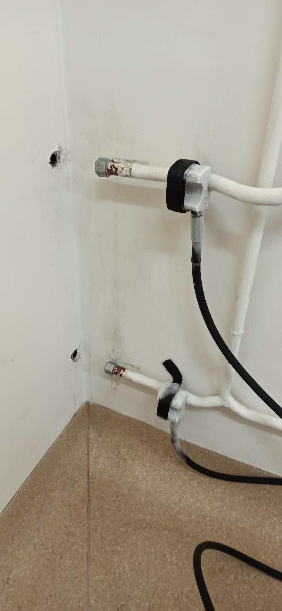Białe rury instalacyjne przy ścianie, zabezpieczone czarną taśmą izolacyjną, widoczne dwa otwory w ścianie po lewej stronie.