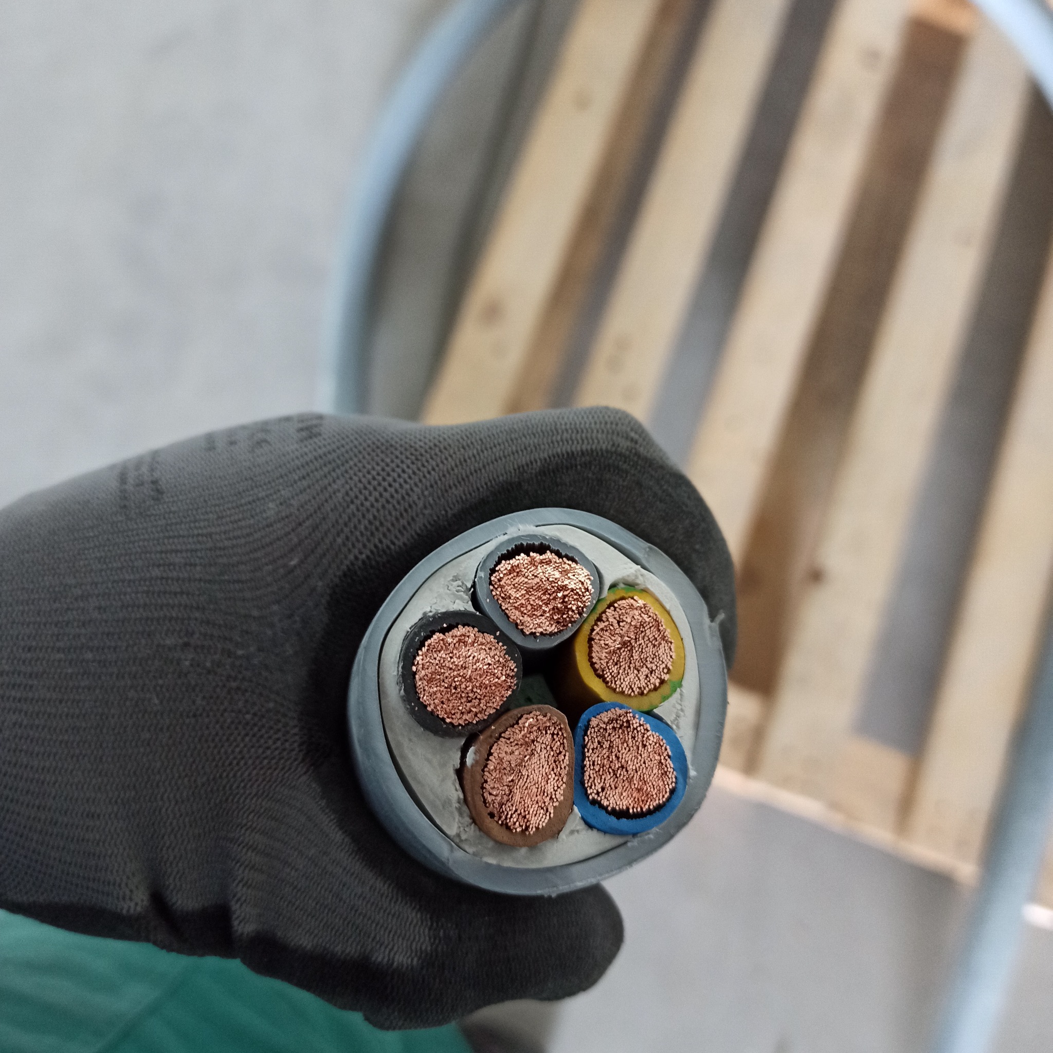 Przekrój grubego kabla elektrycznego z pięcioma żyłami w izolacji o różnych kolorach trzymanego w szarej rękawicy roboczej, w tle drewniana paleta.