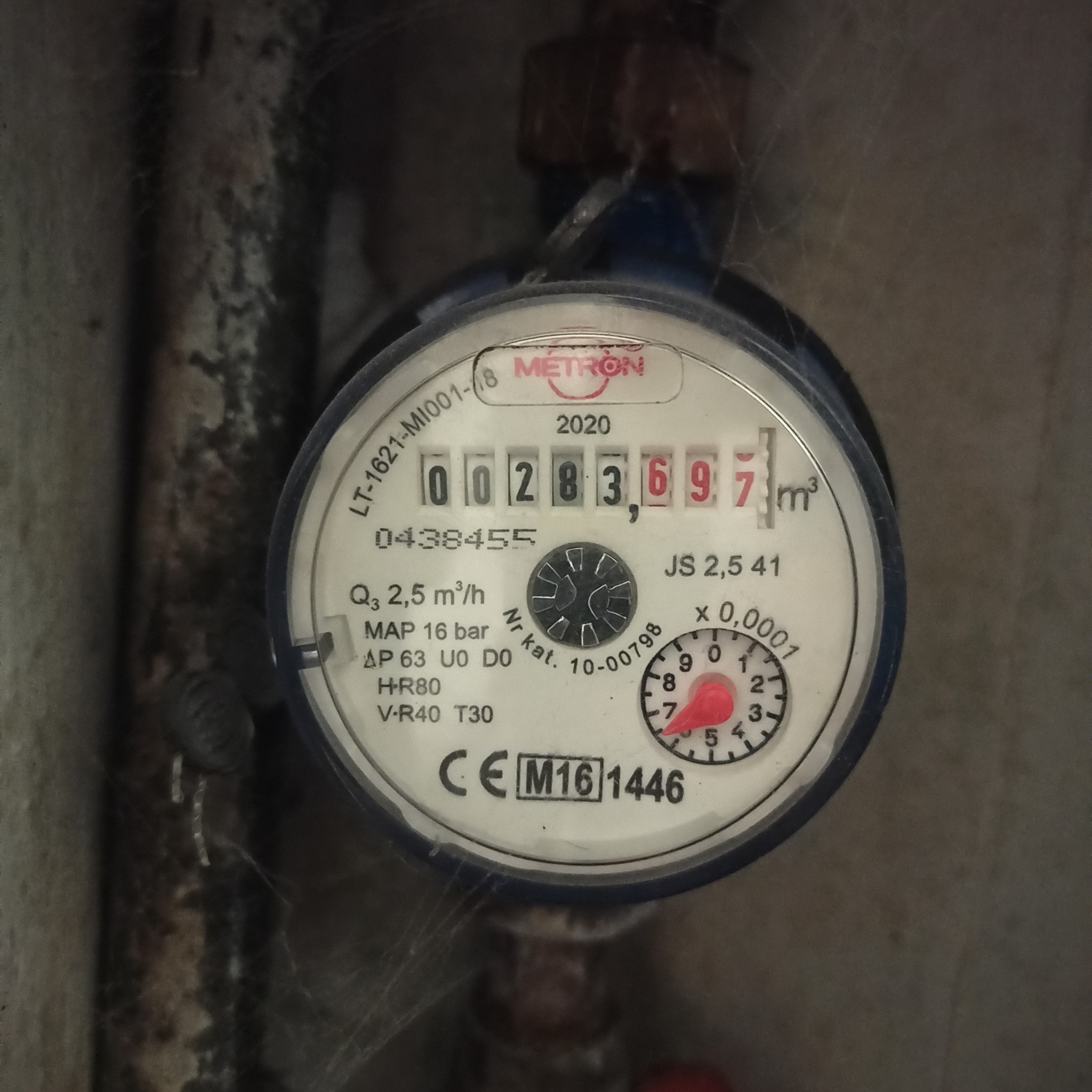 Licznik wody METRON z widocznym wskazaniem 283,697 m3, zamontowany przy ścianie, z widocznymi pajęczynami.