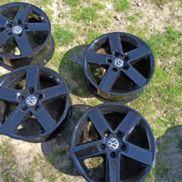 Cztery czarne, aluminiowe felgi samochodowe z logo VW, leżące na trawie w słoneczny dzień.