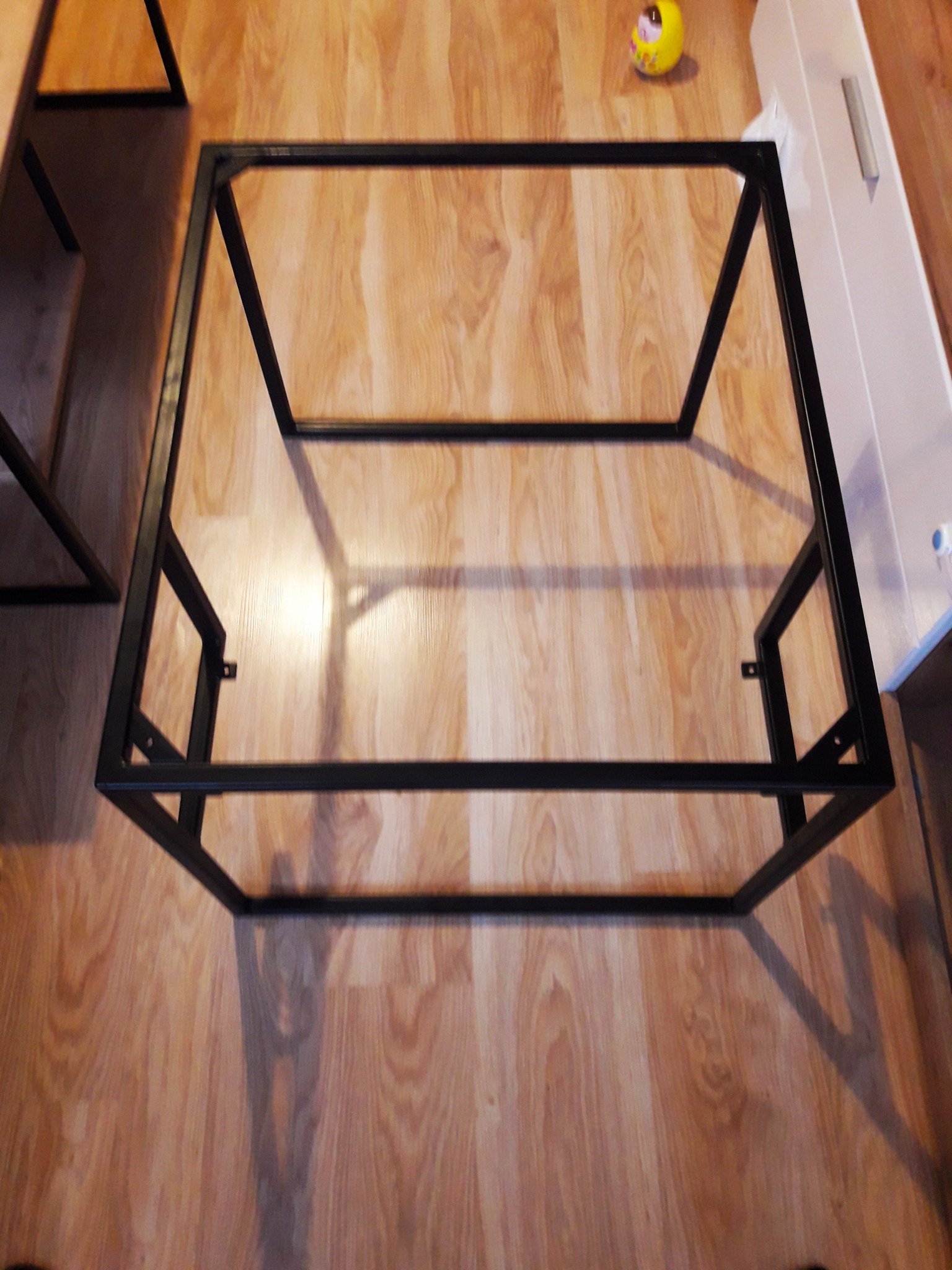 Czarna, metalowa konstrukcja stolika kawowego na drewnianej podłodze, widoczne spawy i mocowania, obok biała szafka i żółta zabawka.