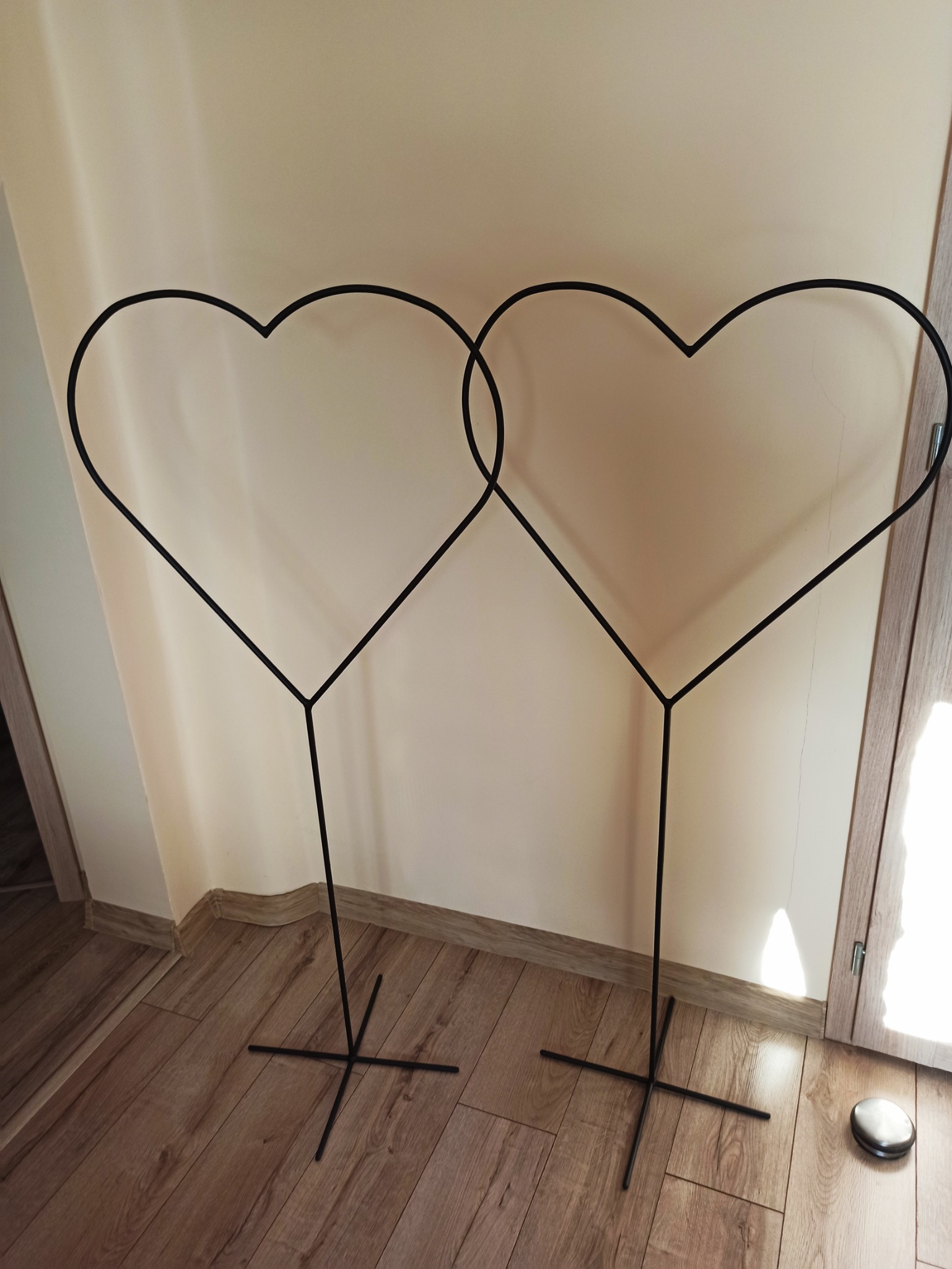 Dwa czarne, metalowe stelaże w kształcie serc, stojące na drewnianej podłodze przy ścianie.