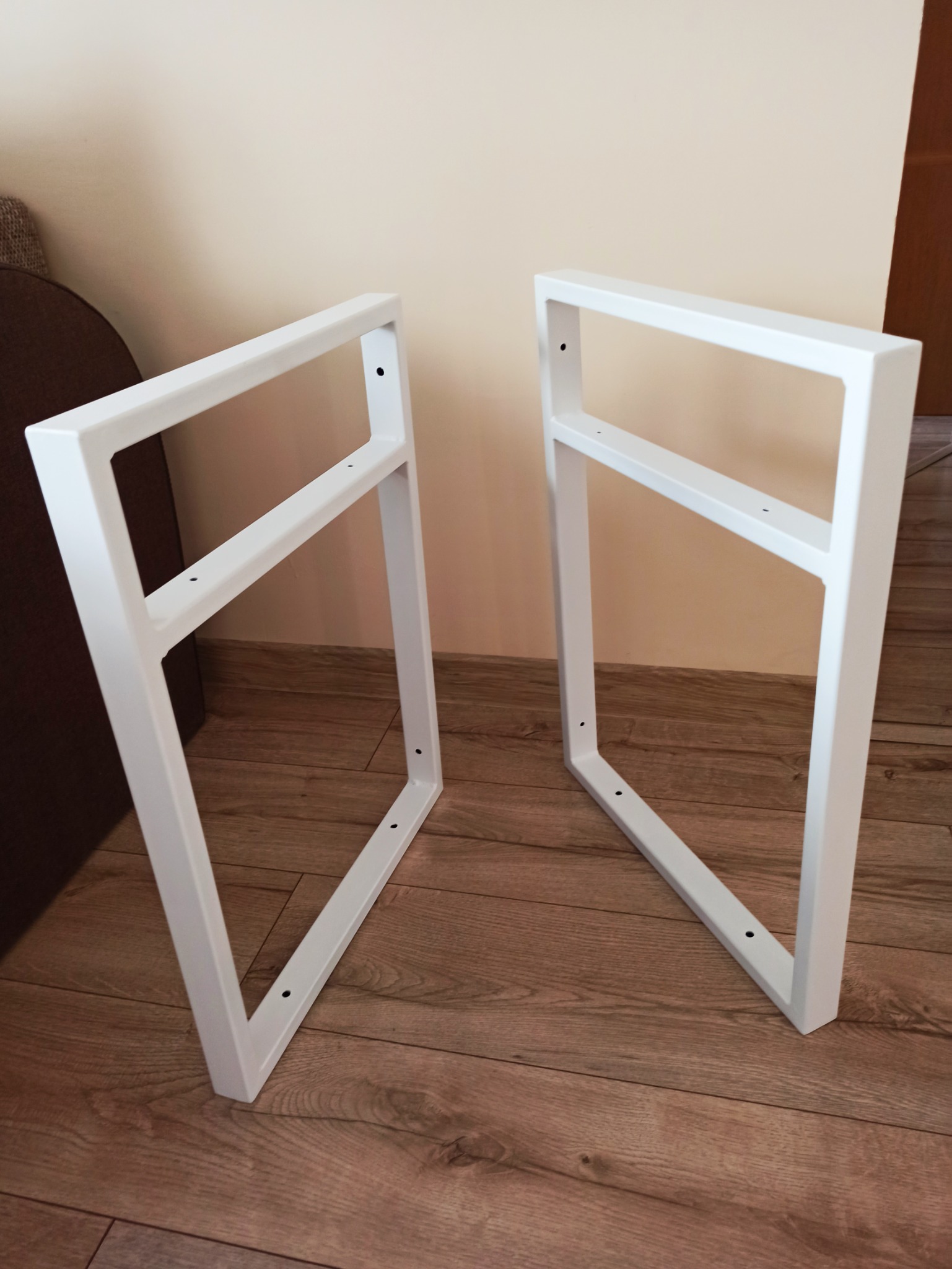 Dwie białe, kwadratowe ramy metalowe z otworami na śruby, prawdopodobnie nogi do stołu, wykonane w Sierpcu, leżą na drewnianej podłodze.