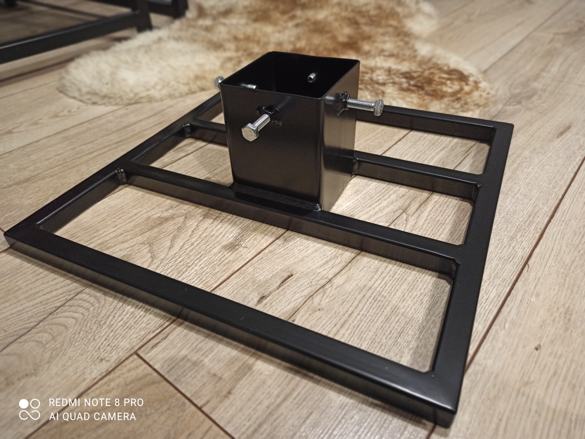 Czarny, spawany, metalowy stojak na choinkę z regulowanymi śrubami, umieszczony na panelach podłogowych z efektem drewna.