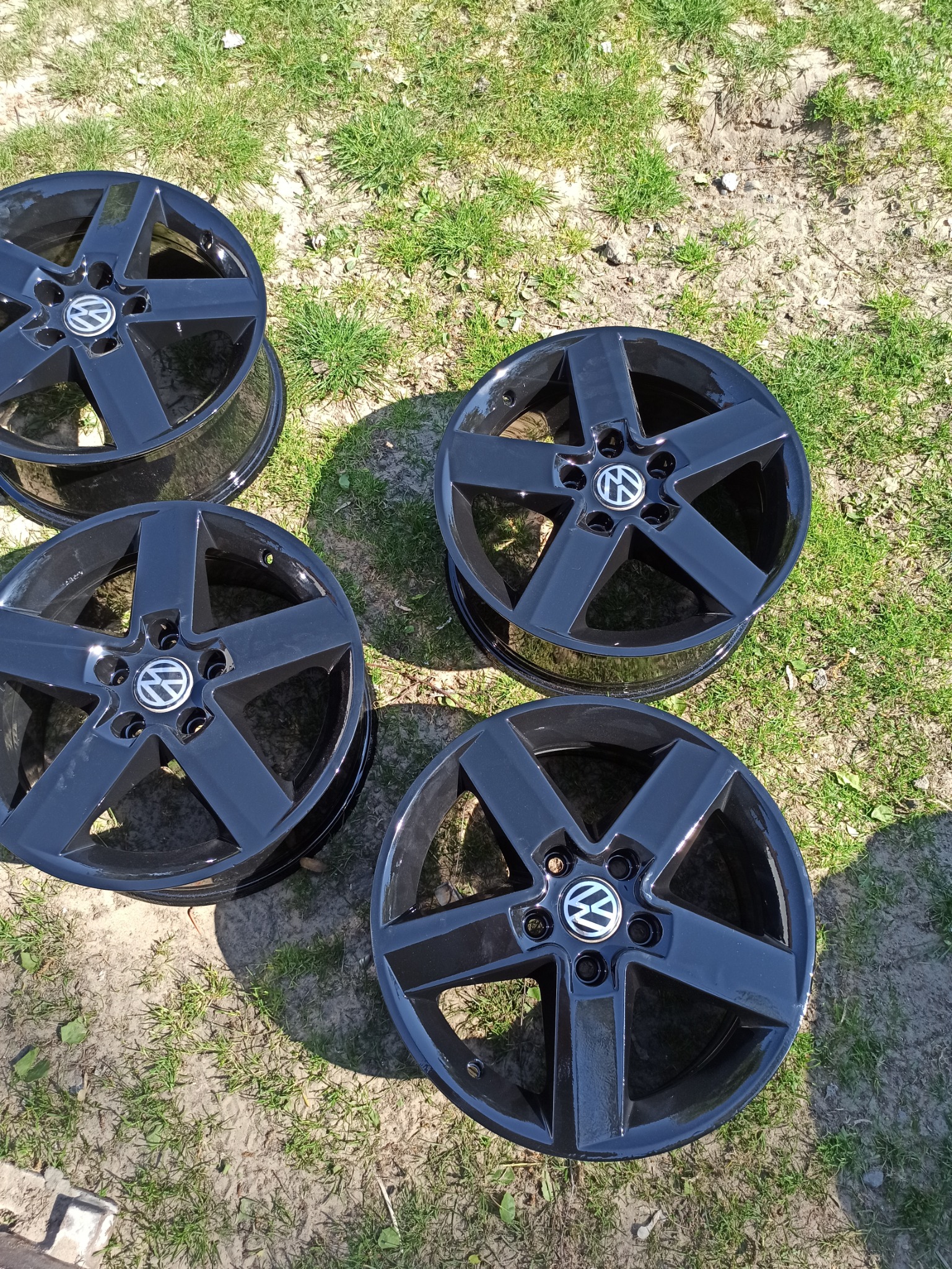 Cztery czarne, aluminiowe felgi samochodowe z logo VW, leżące na trawie w słoneczny dzień.