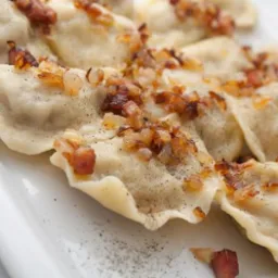 Pierogi okraszone skwarkami i cebulką, ułożone na białym talerzu, posypane pieprzem.