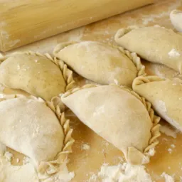 Surowe pierogi ułożone na drewnianym blacie posypanym mąką, obok leży drewniany wałek.