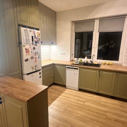 Meble Rostkowski - Nowoczesna kuchnia w odcieniach zieleni z drewnianymi blatami, białymi płytkami na ścianie i lodówką ozdobioną magnesami, widok na okno z odbiciem sylwetek.