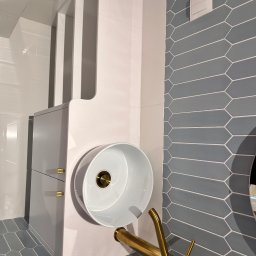 Meble Rostkowski - Nowoczesna łazienka z umywalką nablatową w kształcie walca, złotą baterią i szarą mozaiką w romby na ścianie, szafka pod umywalką z dwoma szufladami i złotymi uchwytami.