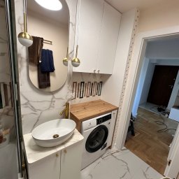 Meble Rostkowski - Nowoczesna łazienka z umywalką nablatową, złotą armaturą, lustrem w kształcie łuku i pralką pod drewnianym blatem, całość utrzymana w bieli i marmurze.