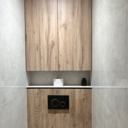 Meble Rostkowski - Drewniana szafka wisząca nad zabudowaną toaletą w łazience wykończonej szarymi płytkami imitującymi kamień.