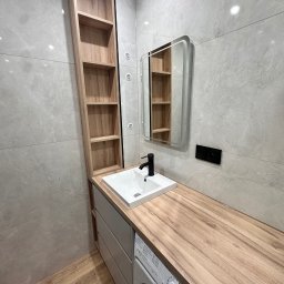 Meble Rostkowski - Nowoczesna łazienka z drewnianym blatem nad pralką, białą umywalką, czarną baterią i wysoką, drewnianą półką w zabudowie, uzupełniona o lustro z podświetleniem.