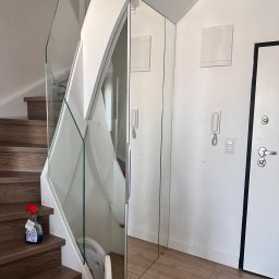 Meble Rostkowski - Nowoczesne schody z drewnianymi stopniami i szklaną balustradą, przylegające do lustrzanej szafy w przedpokoju z drewnianą podłogą i białą ścianą z domofonem.