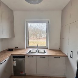 Meble Rostkowski - Jasna kuchnia z drewnianym blatem, zlewozmywakiem dwukomorowym pod oknem i zabudowaną zmywarką, widok na drzewa za oknem.