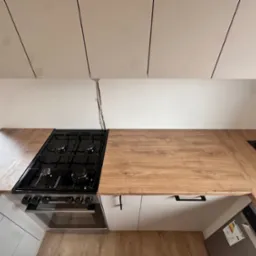 Nowoczesna kuchnia z blatem drewnianym, czarną płytą gazową i białymi szafkami, widok z góry na ukośnym poddaszu, widoczne gniazdka w blacie