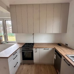 Meble Rostkowski - Jasna kuchnia z drewnianym blatem i białymi szafkami, widoczny fragment okna i zabudowy kuchennej z piekarnikiem i płytą gazową.