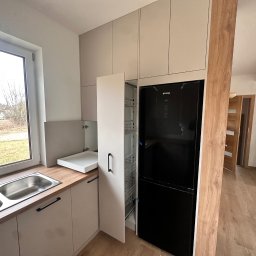 Meble Rostkowski - Jasna kuchnia z drewnianym blatem, stalowym zlewem i otwartą szafką cargo obok czarnej lodówki, widok na schody w tle.