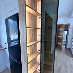 Meble Rostkowski - Wysoka szafa wnękowa z oświetlonymi półkami z drewna, przeszklone drzwiczki z czarną ramą, widok na wnętrze domu.